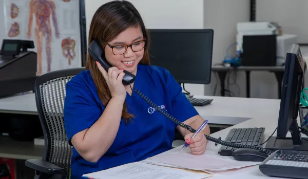 A medical office administrator sits in front of her computer and answers a phone call from an unseen patient.