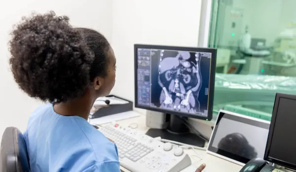 Radiologic technologist analyzing abdominal CT scan on monitor in a medical imaging control room.