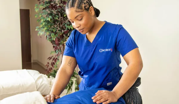 A professional massage therapist wearing blue scrubs with a Concorde logo is performing a leg massage on a client in a bright treatment room.