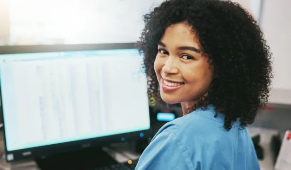 Smiling woman in blue scrubs working at a computer, representing Medical Office Administration Certificate Program skills.