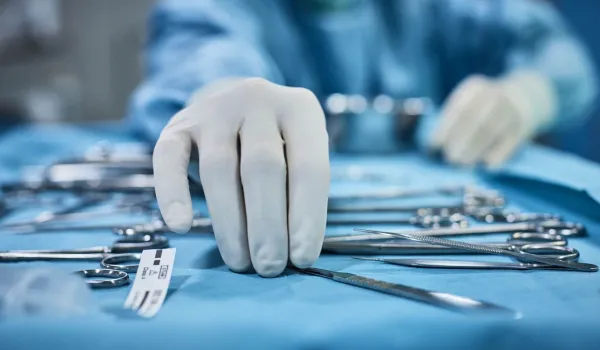 A close-up view of a gloved hand, representing a surgical technologist reaching for a scalpel among a spread of various sterile surgical instruments on a blue instrument tray.