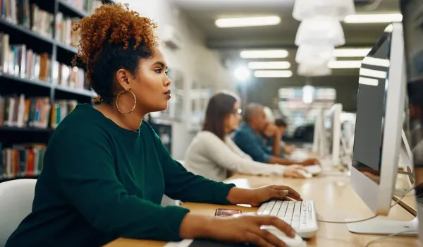 A focused woman with curly hair sits at a computer in a library or lab, working on the coursework that may be part of her healthcare administration degree prerequisites.