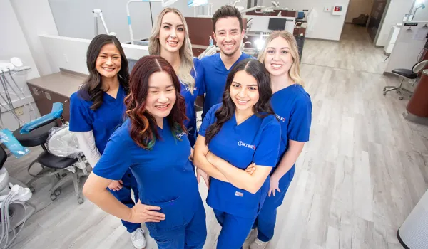 A group of smiling Concorde dental hygiene alumni stand together in blue scrubs within a Concorde dental training facility after completing their dental hygienist requirements.