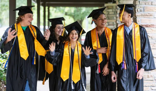 Five high school seniors wearing black graduation caps and gowns with gold stoles smile and celebrate together, representing recipients of a high school senior grant.