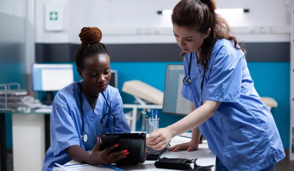A medical assistant and a nurse in blue scrubs collaborate in a modern clinic, reviewing patient data on a digital tablet to illustrate the differences between their clinical and administrative roles.