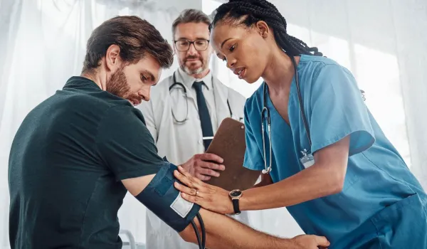 A medical assistant in blue scrubs applies a blood pressure cuff to a patient's arm while being supervised by a senior doctor in a clinical setting.