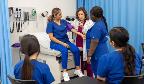 A healthcare professional and educator demonstrates how to take a patient's blood pressure using a student, while other students in the class watch