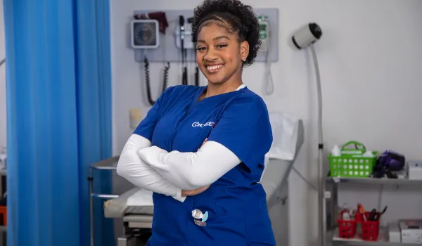 A female medical assistant wearing blue scrubs stands with arms crossed and smiling in a hospital setting