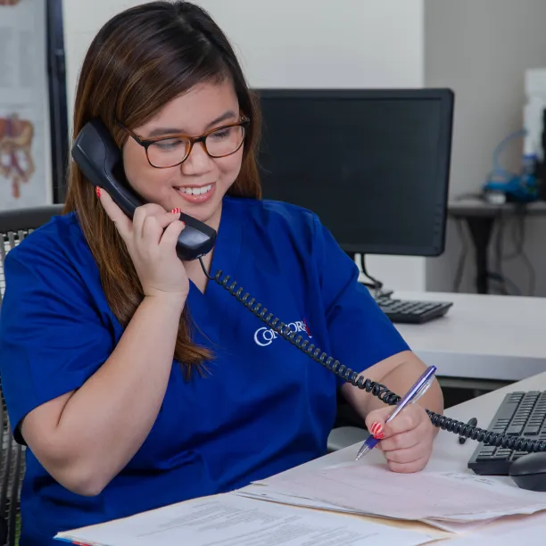 A medical office administrator sits in front of her computer and answers a phone call from an unseen patient.