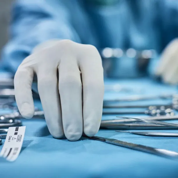 A close-up view of a gloved hand, representing a surgical technologist reaching for a scalpel among a spread of various sterile surgical instruments on a blue instrument tray.
