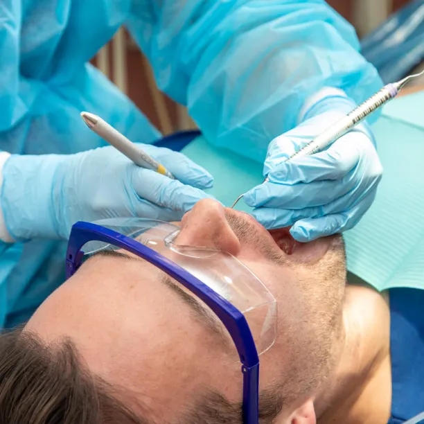 A close-up view of a dental hygienist wearing a blue gown and gloves using dental tools to work on a male patient who is wearing protective eyewear.
