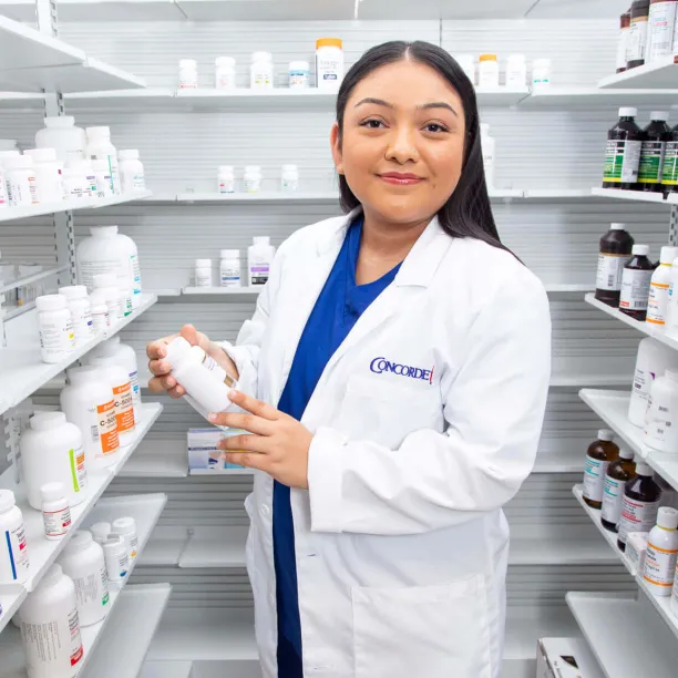 A smiling female pharmacy technician in a white lab coat stands in an aisle between shelves full of medication, holding a bottle to check the label.