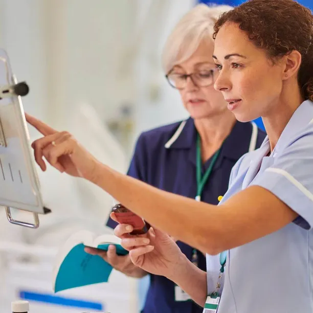 Two female nurses in professional uniforms review medical data on a wall-mounted digital terminal while one holds a medication bottle and a reference book.