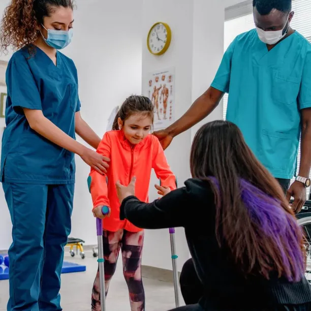 A pediatric physical therapist assistant supports a young girl during a therapeutic exercise session in a brightly colored clinic.