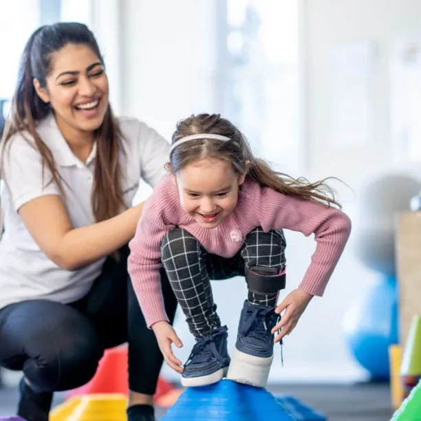 A young child balances on a large blue therapy ball while being guided through stability exercises by a pediatric physical therapist assistant.