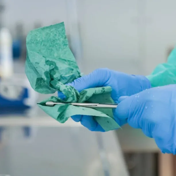 A person wearing a blue protective gown and surgical gloves carefully wipes and cleans a small surgical instrument during the decontamination phase of sterile processing.