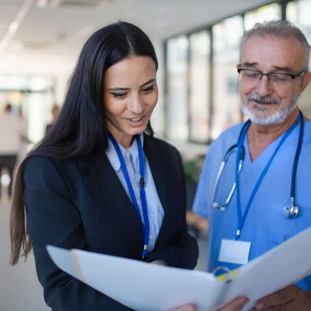 A female healthcare administrator in a black suit reviews medical documents in a binder with a senior male doctor in blue scrubs.