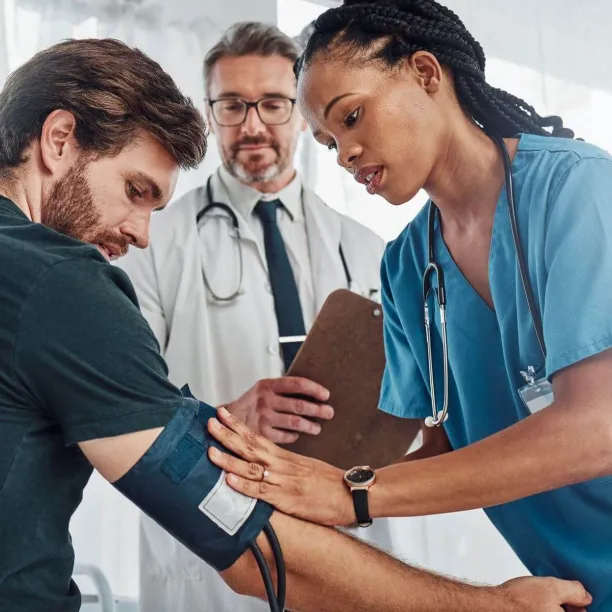A medical assistant in blue scrubs applies a blood pressure cuff to a patient's arm while being supervised by a senior doctor in a clinical setting.