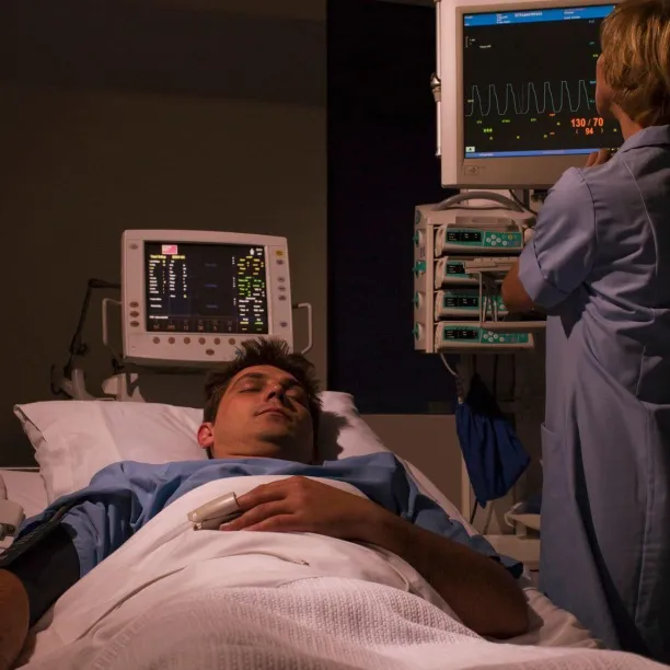 A sleep technologist monitors real-time physiological data and vital signs on multiple screens while a patient sleeps in a darkened room.