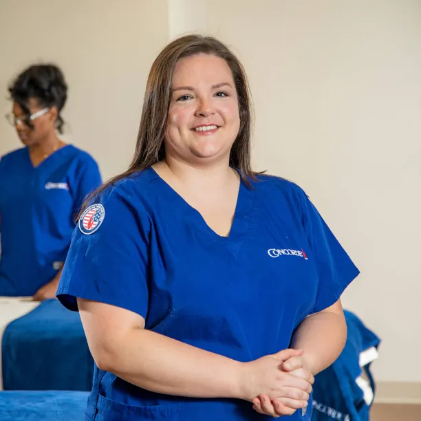 A smiling Concorde massage therapy student wearing blue scrubs with a military-themed patch stands in a healthcare training lab, illustrating how to use military tuition assistance for career training.
