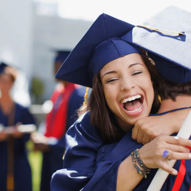 Female graduate in blue cap and gown holding diploma hugs another graduate with big smile on her face.