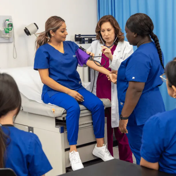 A healthcare professional and educator demonstrates how to take a patient's blood pressure using a student, while other students in the class watch
