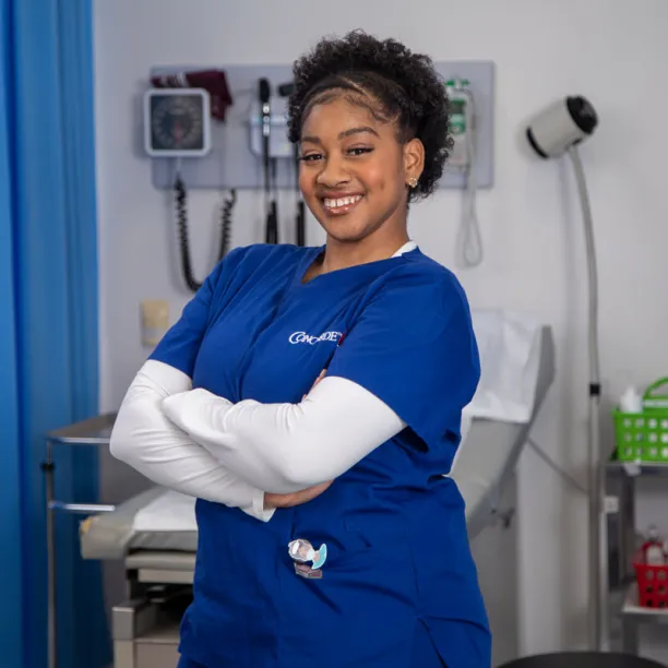 A female medical assistant wearing blue scrubs stands with arms crossed and smiling in a hospital setting