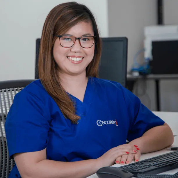 female medical office adminstrator in blue scrubs sits at computer and smiles for camera