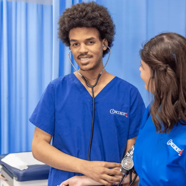 A male medical assistant wearing blue scrubs and stethescope works with patient and smiles for camera