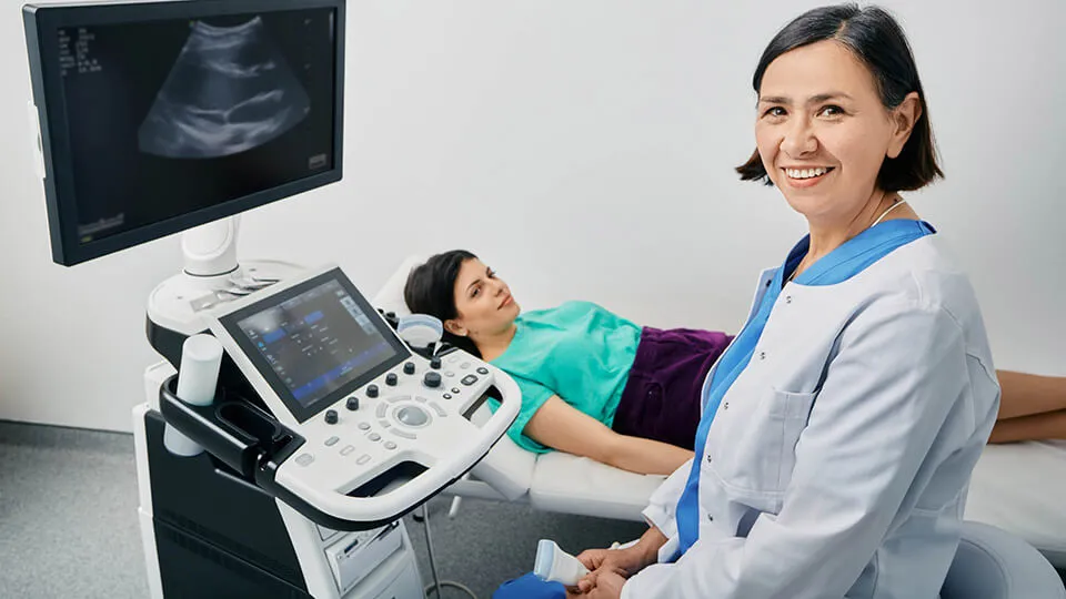 A sonographer smiles at the camera while her patient lies on a bed next to the scanning equipment