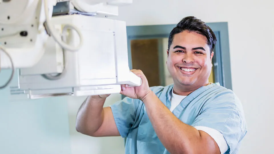 A man wearing scrubs smiles as he positions imaging equipment in a medical facility. A man wearing scrubs smiles as he positions imaging equipment in a medical facility.