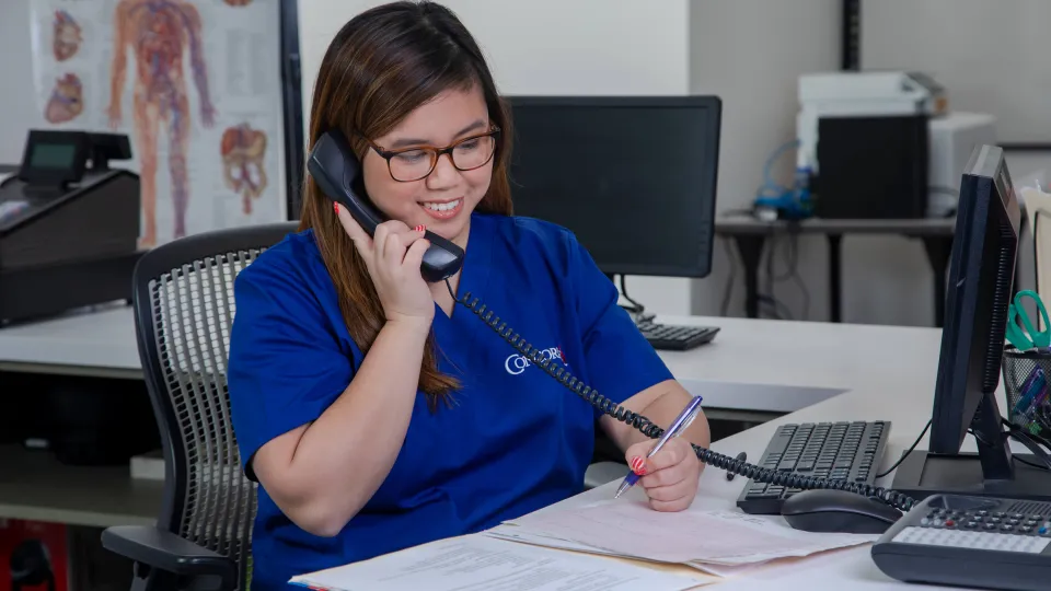 A medical office administrator sits in front of her computer and answers a phone call from an unseen patient. A medical office administrator sits in front of her computer and answers a phone call from an unseen patient.