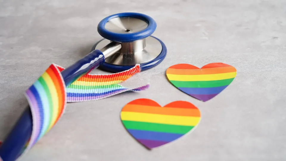 Stethoscope wrapped around rainbow pride ribbon with rainbow pride flag and pride flag hear cutouts on table