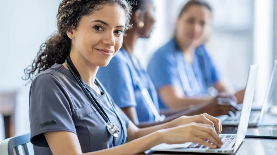 Smiling medical assistant student with stethoscope using laptop in class, representing medical assistant certification training.