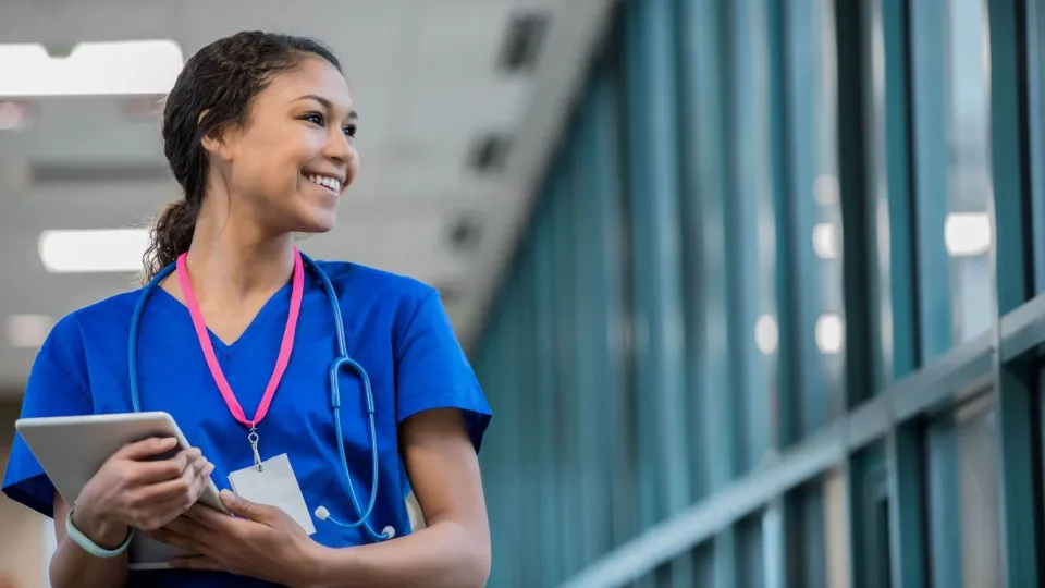Smiling travel nurse in scrubs holding a tablet in a hospital hallway, representing confidence and readiness for a traveling nurse career Smiling travel nurse in scrubs holding a tablet in a hospital hallway, representing confidence and readiness for a traveling nurse career