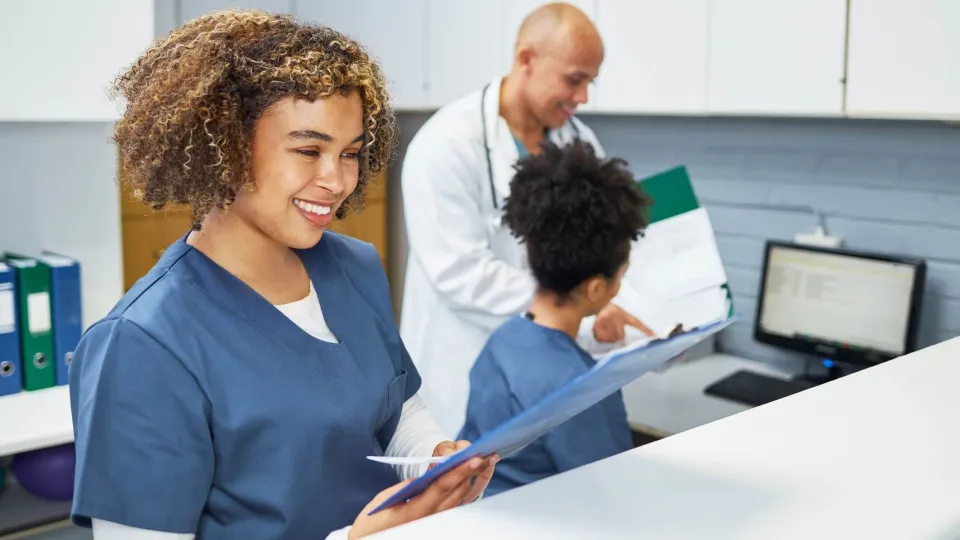 A smiling medical assistant in blue scrubs stands at a reception counter reviewing a patient chart while a doctor and another medical assistant discuss data on a computer screen in the background. A smiling medical assistant in blue scrubs stands at a reception counter reviewing a patient chart while a doctor and another medical assistant discuss data on a computer screen in the background.