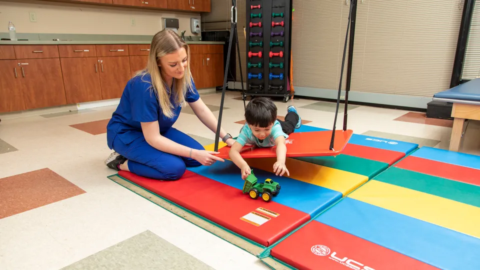 Occupational therapy assistant guiding a child through sensory motor play using colorful mats and a toy tractor during a pediatric therapy session. Occupational therapy assistant guiding a child through sensory motor play using colorful mats and a toy tractor during a pediatric therapy session.