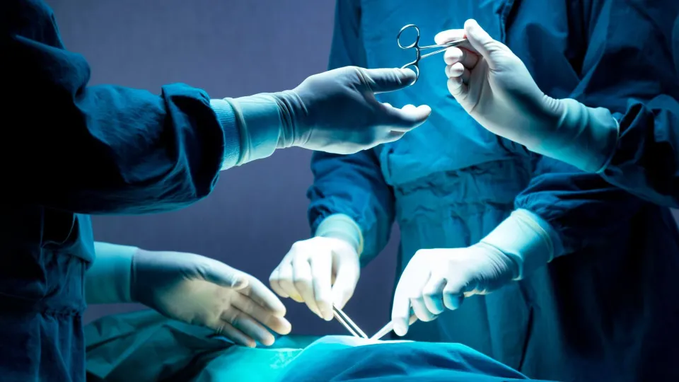 A surgical team in dark blue gowns works together under dim lighting in an operating room, with one gloved hand passing surgical scissors to another member of the team over the draped patient.