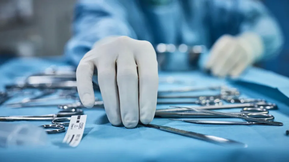 A close-up view of a gloved hand, representing a surgical technologist reaching for a scalpel among a spread of various sterile surgical instruments on a blue instrument tray. A close-up view of a gloved hand, representing a surgical technologist reaching for a scalpel among a spread of various sterile surgical instruments on a blue instrument tray.