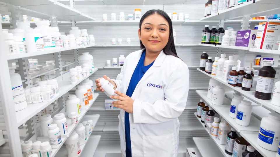 A smiling female pharmacy technician in a white lab coat stands in an aisle between shelves full of medication, holding a bottle to check the label. A smiling female pharmacy technician in a white lab coat stands in an aisle between shelves full of medication, holding a bottle to check the label.