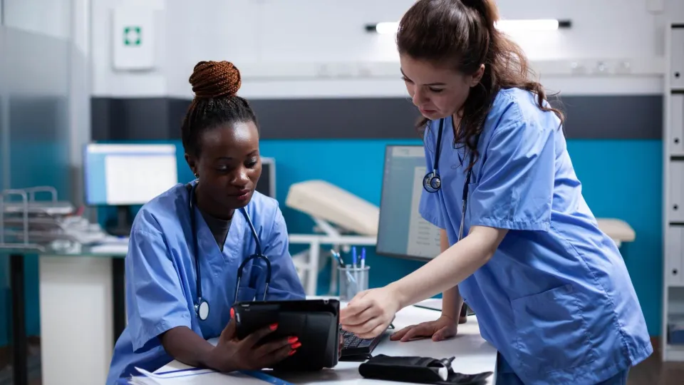 A medical assistant and a nurse in blue scrubs collaborate in a modern clinic, reviewing patient data on a digital tablet to illustrate the differences between their clinical and administrative roles.