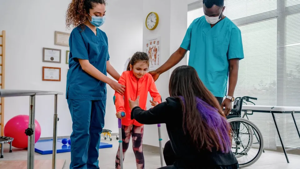 A pediatric physical therapist assistant supports a young girl during a therapeutic exercise session in a brightly colored clinic.