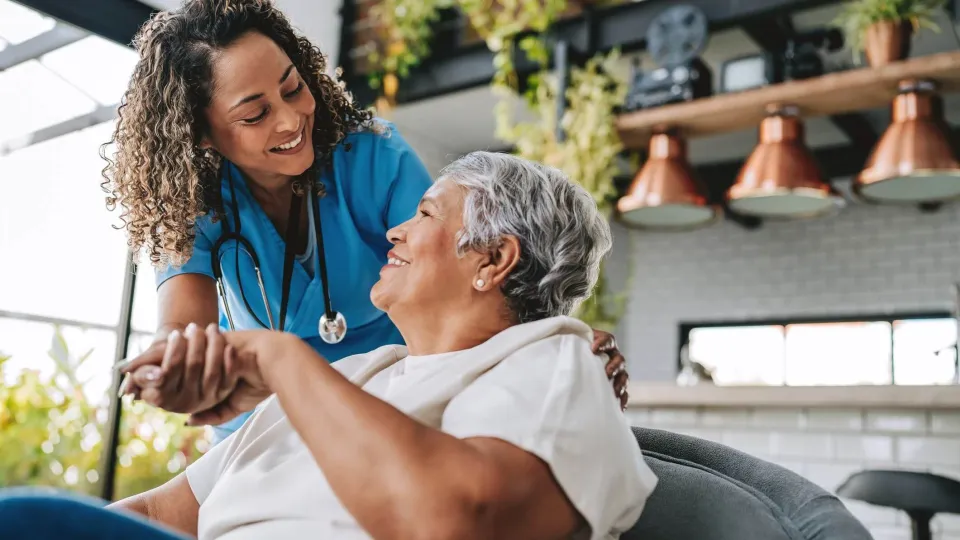 A smiling nurse holds the hand of an elderly patient in a warm, bright room, illustrating the rewarding patient-care aspect of changing your career to nursing.