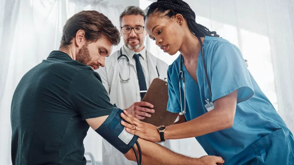 A medical assistant in blue scrubs applies a blood pressure cuff to a patient's arm while being supervised by a senior doctor in a clinical setting.