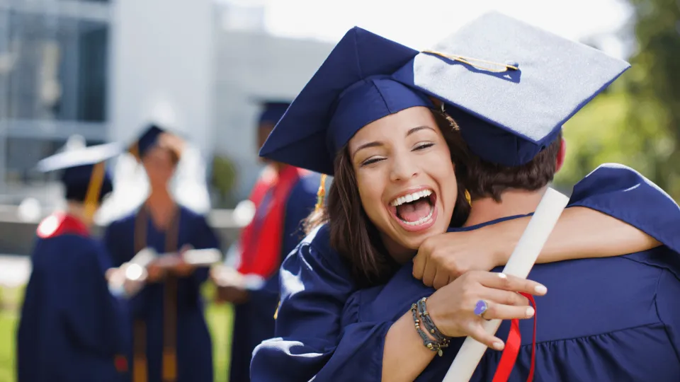 Female graduate in blue cap and gown holding diploma hugs another graduate with big smile on her face.