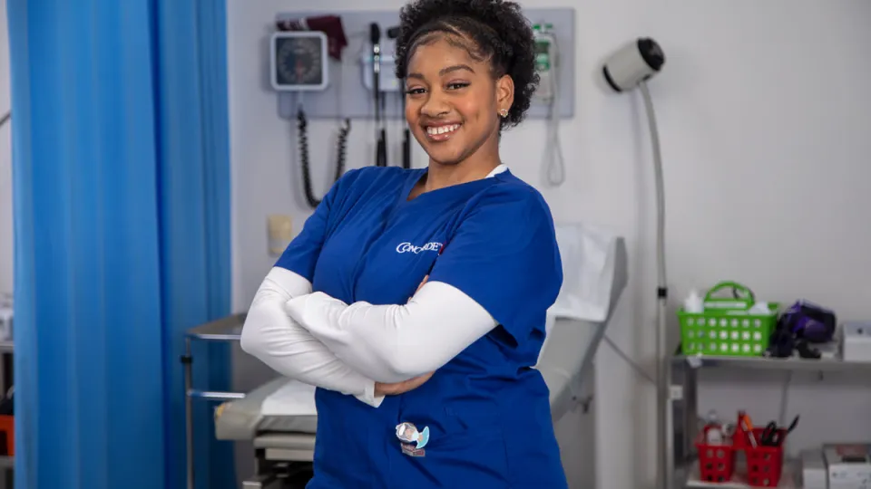 A female medical assistant wearing blue scrubs stands with arms crossed and smiling in a hospital setting