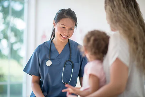 A vocational nurse smiles at a baby being held by its mother