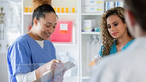 Pharmacy tech in scrubs holding clipboard beside colleagues