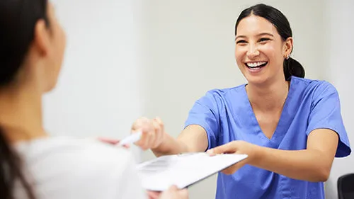 A smiling medical assistant hands a form on a clipboard to a patient to fill out