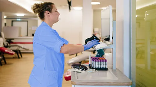 A phlebotomist inputs data into a computer while vials of blood samples and a blood bag sit on the table beside her.